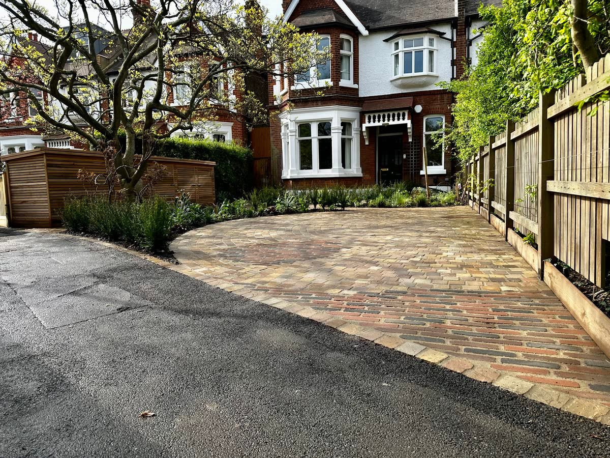 Reclaimed stone sett driveway laid in a fan pattern, edged with a planted border and a slatted timber bin store, in front of a bay-fronted Edwardian semi